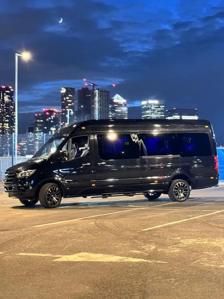 A sleek black Mercedes Monaco Sprinter luxury van parked at night with the illuminated Canary Wharf skyline in the background. The vehicle's glossy finish reflects the city lights, while the ambient blue interior lighting adds to its premium appeal. A crescent moon shines above, enhancing the sophisticated urban setting.